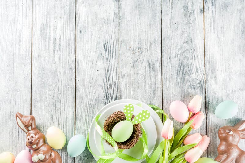 Easter holiday table setting with rabbits and eggs, plates with colorful eggs and chocolate bunny rabbits, festive ribbon, wooden background top view copy space