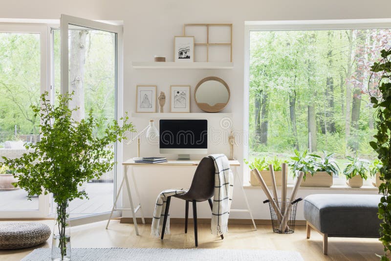 Echte foto van wit woonkamerbinnenland met groot venster, glasdeur, verse installaties, houten bureau met modelcomputer en eenvou