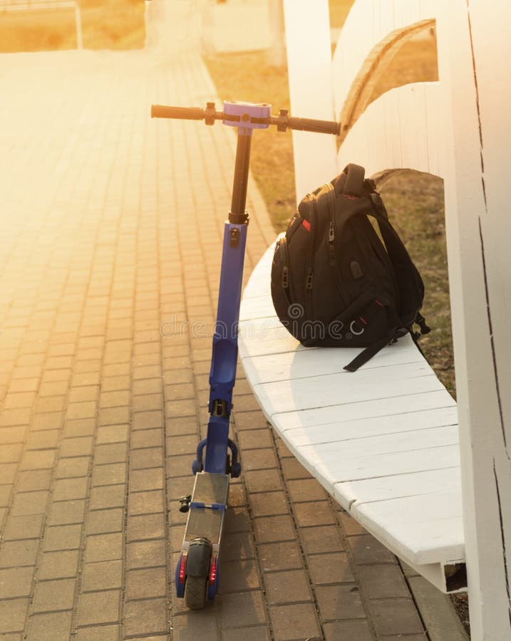 An eco-friendly electric blue scooter stands in a park leaning on a bench, on which lies a black urban backpack. Evening, sunset.