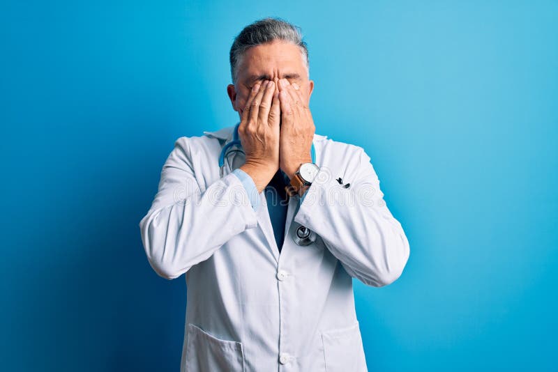 Edad media guapo médico pelirrojo hombre con abrigo y estetoscopio azul frotando los ojos para la fatiga y el dolor de cabeza somn foto de archivo