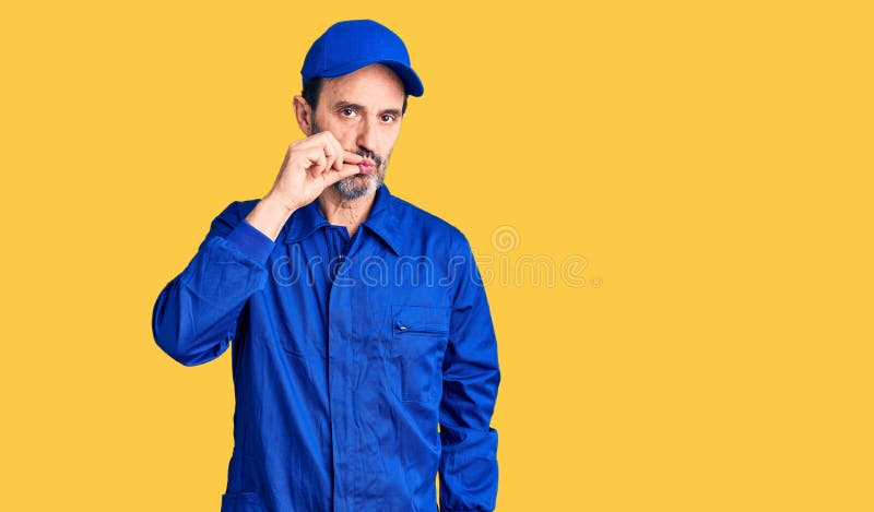 Edad media hombre guapo con uniforme mecánico boca y labios cerrados como cremallera con los dedos foto de archivo libre de regalías