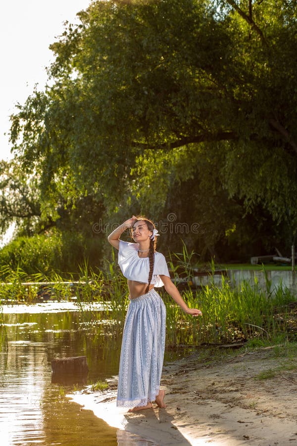 On the edge of the shore near the river stands a girl in a white dress.