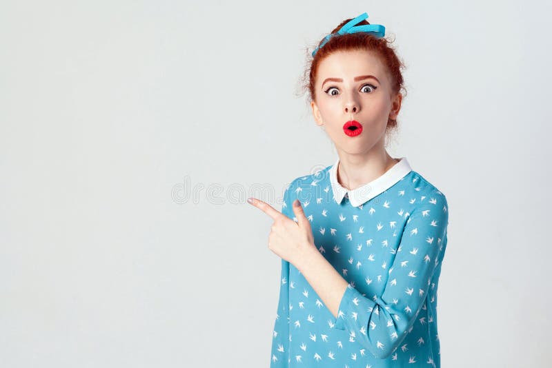 Human face expressions, emotions and feelings. Astonished and shocked young redhead girl in blue dress, pointing at blank studio wall, surprised with sale prices, keeping her mouth wide open. Isolated studio shot on gray background