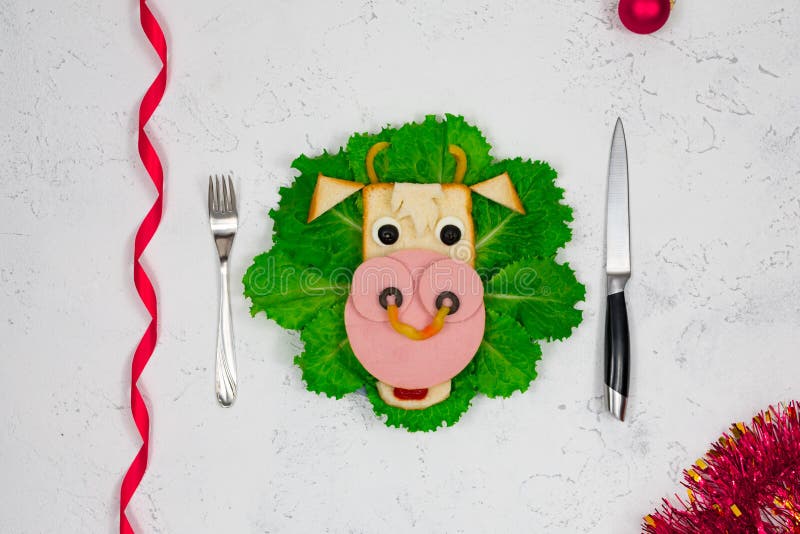 An edible symbol of 2021. Creative children`s sandwich in the form of a bull, a cow on a plate. Cute sandwich in the shape of a cow with sausage, olives, cheese, on lettuce leaves. Table, tinsel. New Year`s menu.
