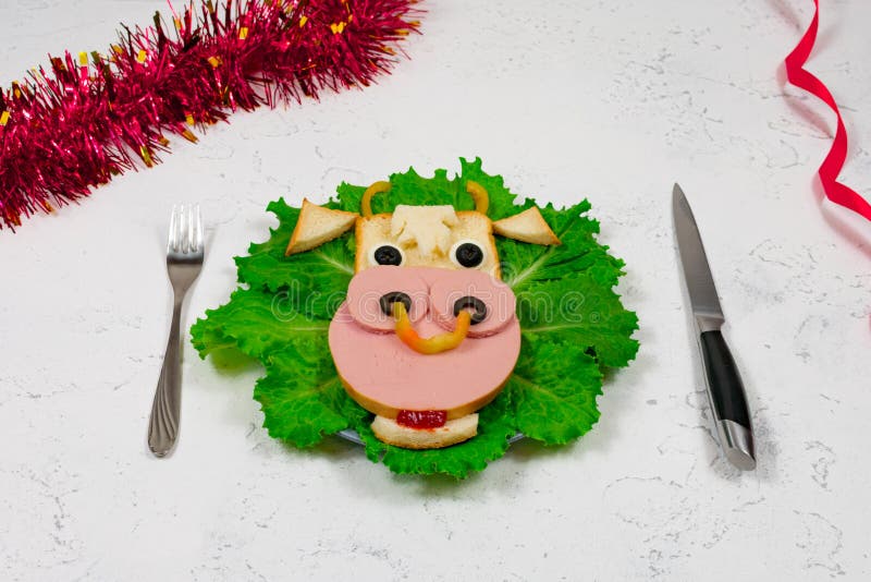 An edible symbol of 2021. Creative children`s sandwich in the form of a bull, a cow on a plate. Cute sandwich in the shape of a cow with sausage, olives, cheese, on lettuce leaves. Table, tinsel. New Year`s menu.