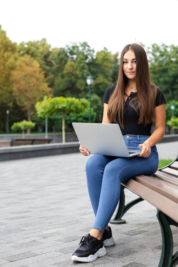 Education, business, technology, and Internet concept — Smiling European dark-haired girl in modern clothes sitting on a bench in the park in summer and holding a laptop computer