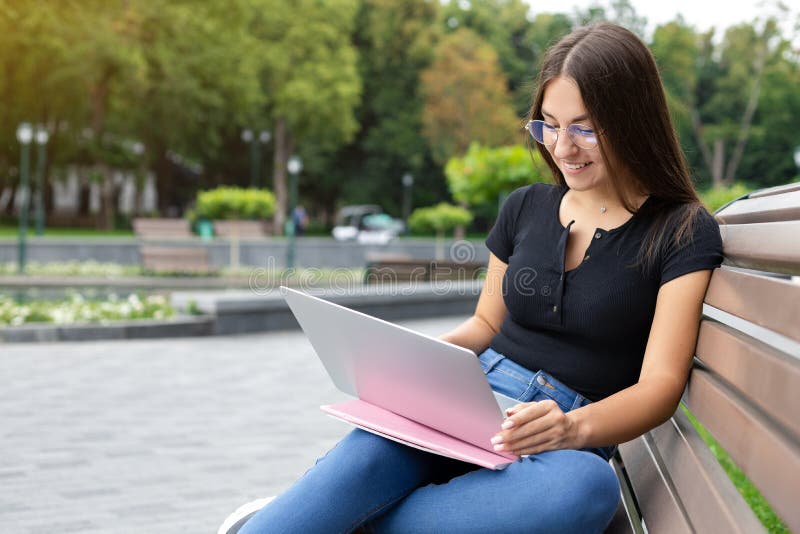 Education, technology, and Internet concept — A smiling European dark-haired student girl sitting on a park bench with her notebooks and looking at her classmates on her laptop while online studying