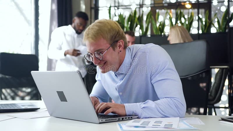 Een aantrekkelijke jonge, blonde zakenman met bril tevreden over de resultaten van zijn werk op de computer, die op kantoor zit