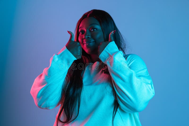 Nice sign, thumbs up. One African beautiful woman with long straight hair isolated on blue studio background in neon light. Concept of human emotions, facial expression. Bodypositive and diversity. Nice sign, thumbs up. One African beautiful woman with long straight hair isolated on blue studio background in neon light. Concept of human emotions, facial expression. Bodypositive and diversity