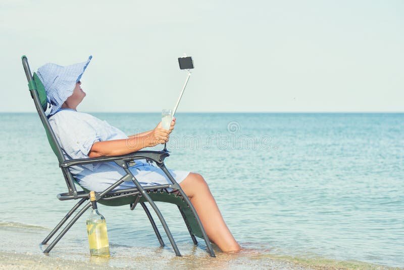 Een bejaarde zit op het strand op een chaise-longue, het drinken wijn en het maken van foto tegen de achtergrond van het overzees stock foto's Een bejaarde zit op het strand op een chaise-longue, het drinken wijn en het maken van foto tegen de achtergrond van het overzees stock foto's