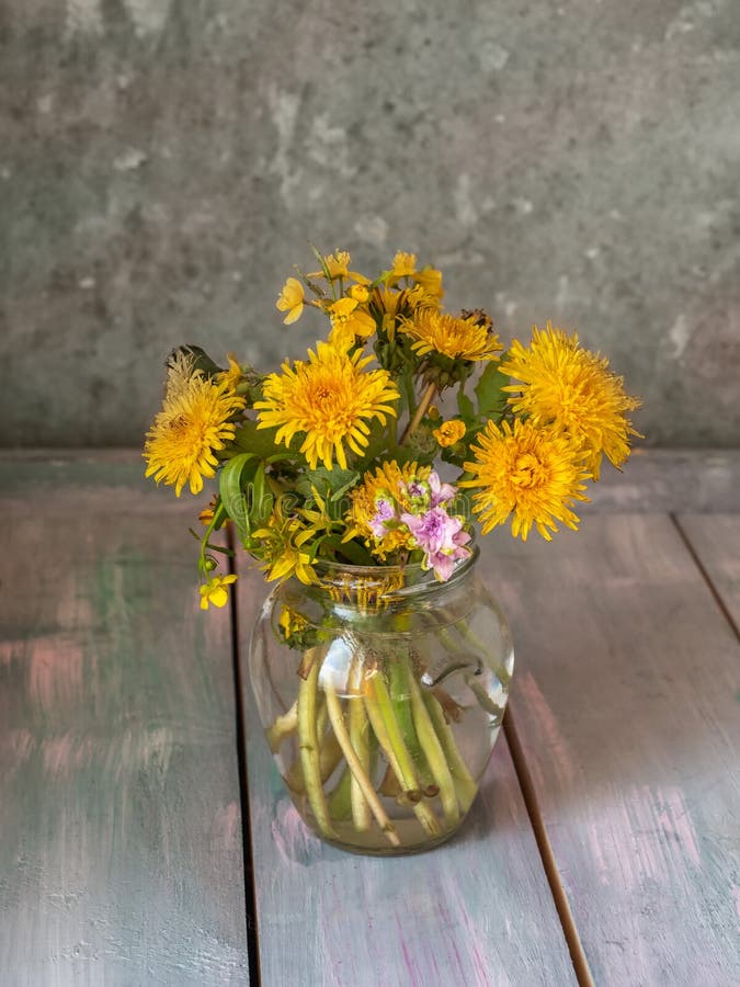 Een boeket van bloeiende paardebloemen en een kleine hyancite in een glaskruik die op een houten lijst, schot bij dichte waaier z stock foto Een boeket van bloeiende paardebloemen en een kleine hyancite in een glaskruik die op een houten lijst, schot bij dichte waaier z stock foto