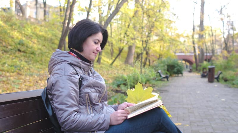 Een brunette vrouw in een bruin jasje die een boek leest terwijl ze op een parkbank zit. herfstzondag