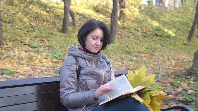 Een brunette vrouw in een bruin jasje die een boek leest terwijl ze op een parkbank zit. herfstzondag