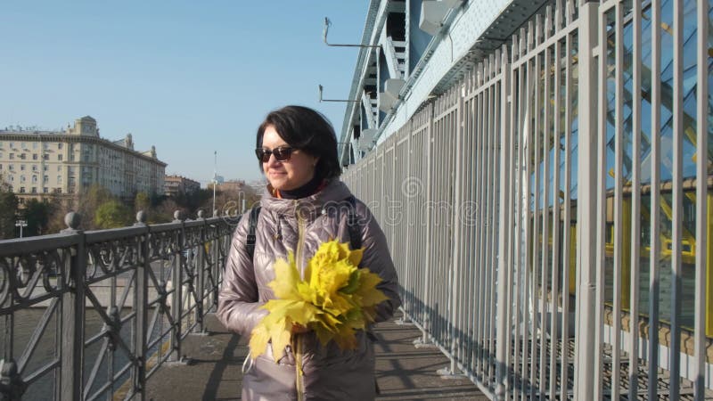 Een brunette vrouw met middelgerijpte bril loopt alleen met een boeket gele bladeren op de brug over de rivier .