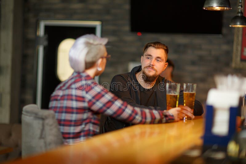 Een donkerbruine kerel met een baard, zit en spreekt in bar met een meisje en drinkt bier binnen stock afbeelding Een donkerbruine kerel met een baard, zit en spreekt in bar met een meisje en drinkt bier binnen stock afbeelding