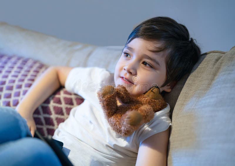 Candid shot of Happy boy with smiling face sitting on sofa watching TV, Positive Child laying on couch playing with dog toy.  Candid shot healhty kid relaxing at home on weekend. Candid shot of Happy boy with smiling face sitting on sofa watching TV, Positive Child laying on couch playing with dog toy.  Candid shot healhty kid relaxing at home on weekend