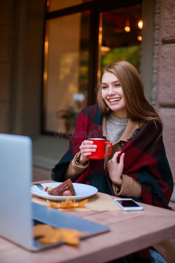 Een gelukkig die meisje, in een deken wordt verpakt, zit in een koffie die aan iemand op laptop spreken, een dessert eten en van stock afbeeldingen Een gelukkig die meisje, in een deken wordt verpakt, zit in een koffie die aan iemand op laptop spreken, een dessert eten en van stock afbeeldingen