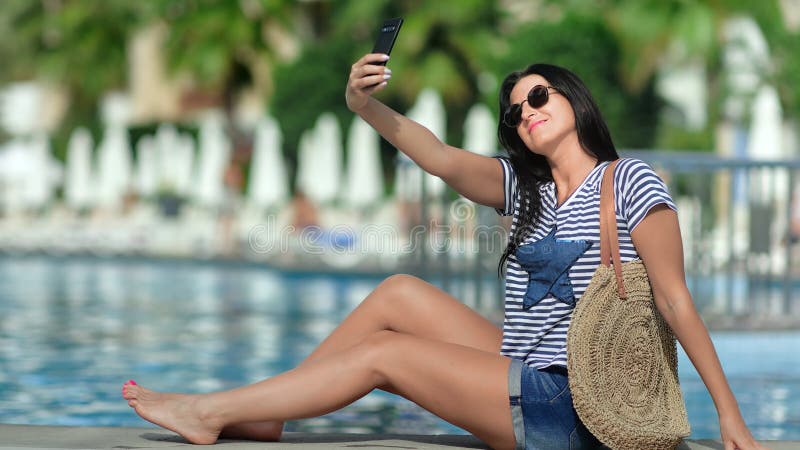 Een glimlach-reisvrouw die zichzelf met behulp van smartphone op de palmachtergrond van het zwembad neemt