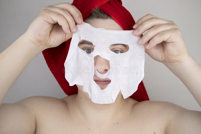 Funny man with a red towel puts a moisturizing fabric mask on his face. The concept of men`s personal care, spa treatments and the use of men`s cosmetics. Funny man with a red towel puts a moisturizing fabric mask on his face. The concept of men`s personal care, spa treatments and the use of men`s cosmetics