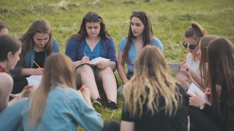 Een groep vrouwelijke studenten zit in een cirkel op een weide voor collectief werk met notebooks.