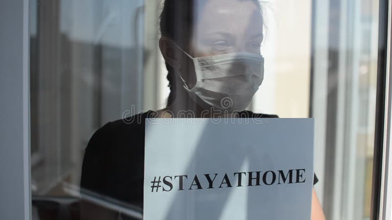 Een jong meisje in een medisch masker staat aan het raam met een teken dat stayathome zegt