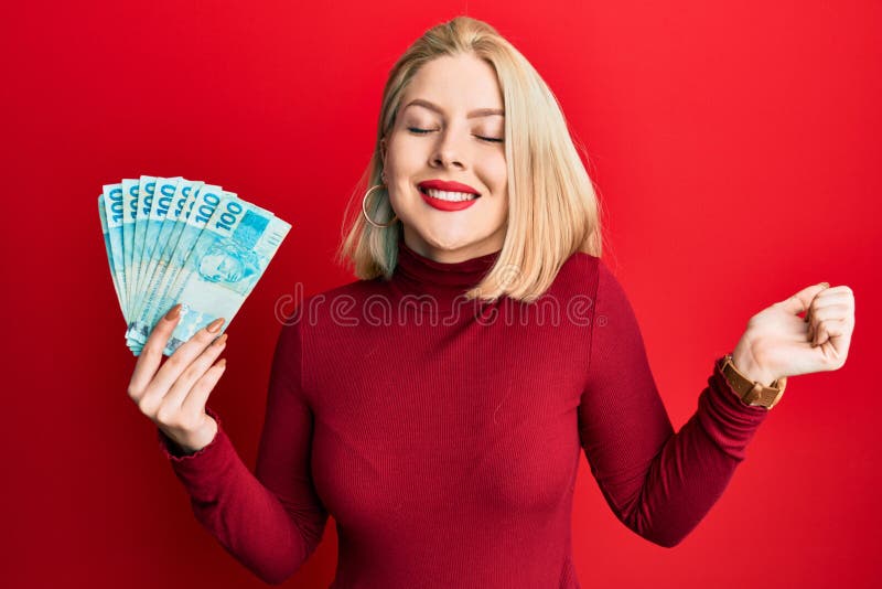 Young blonde woman holding 100 brazilian real banknotes screaming proud, celebrating victory and success very excited with raised arm. Young blonde woman holding 100 brazilian real banknotes screaming proud, celebrating victory and success very excited with raised arm