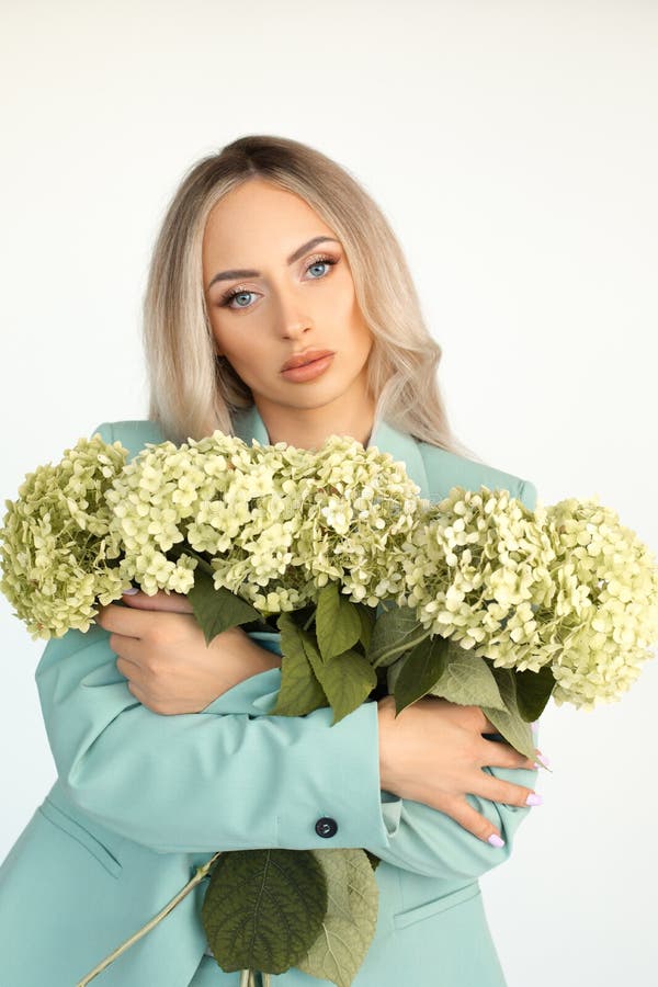 Young beautiful blonde woman with long hair in mint green pantsuit with armful of hydrangeas flowers in arms. Portrait. Young beautiful blonde woman with long hair in mint green pantsuit with armful of hydrangeas flowers in arms. Portrait