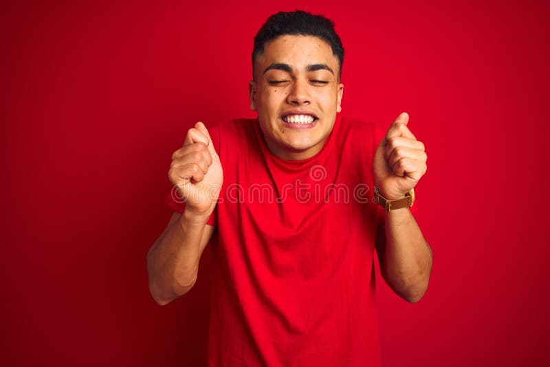 Young brazilian man wearing t-shirt standing over isolated red background excited for success with arms raised and eyes closed celebrating victory smiling. Winner concept. Young brazilian man wearing t-shirt standing over isolated red background excited for success with arms raised and eyes closed celebrating victory smiling. Winner concept