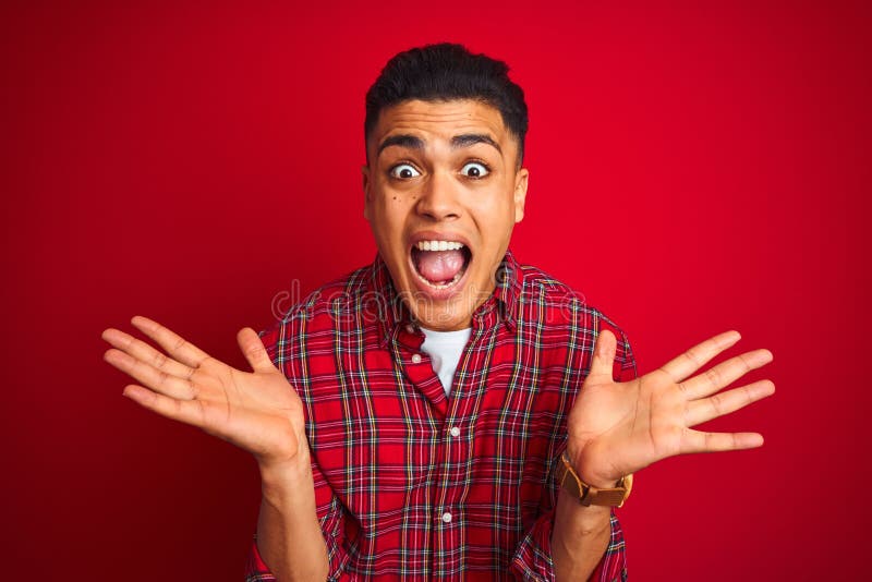 Young brazilian man wearing shirt standing over isolated red background celebrating crazy and amazed for success with arms raised and open eyes screaming excited. Winner concept. Young brazilian man wearing shirt standing over isolated red background celebrating crazy and amazed for success with arms raised and open eyes screaming excited. Winner concept