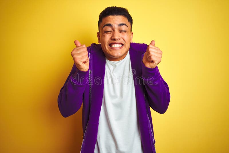 Young brazilian man wearing purple sweatshirt standing over isolated yellow background excited for success with arms raised and eyes closed celebrating victory smiling. Winner concept. Young brazilian man wearing purple sweatshirt standing over isolated yellow background excited for success with arms raised and eyes closed celebrating victory smiling. Winner concept