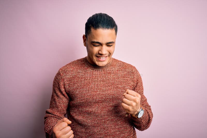 Young brazilian man wearing casual sweater standing over isolated pink background excited for success with arms raised and eyes closed celebrating victory smiling. Winner concept. Young brazilian man wearing casual sweater standing over isolated pink background excited for success with arms raised and eyes closed celebrating victory smiling. Winner concept