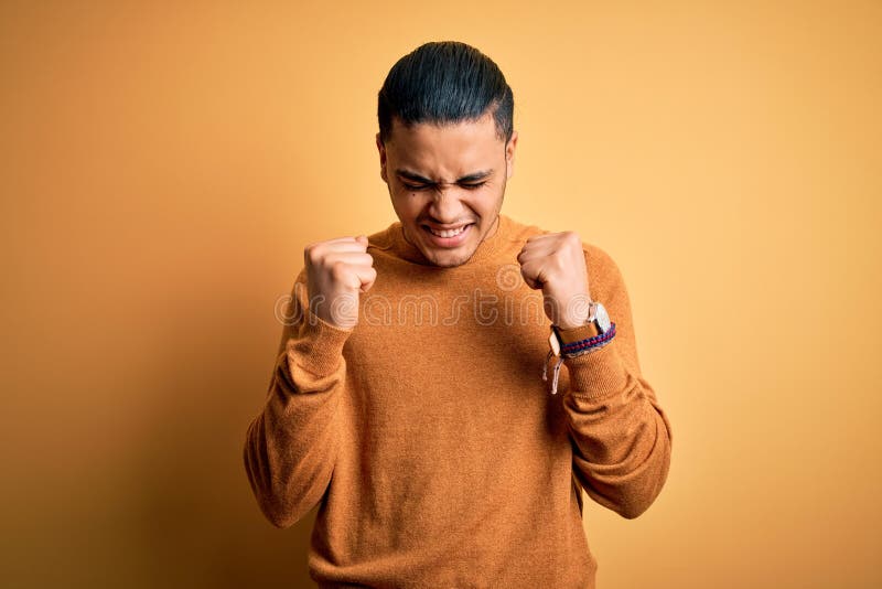 Young brazilian man wearing casual sweater standing over isolated yellow background excited for success with arms raised and eyes closed celebrating victory smiling. Winner concept. Young brazilian man wearing casual sweater standing over isolated yellow background excited for success with arms raised and eyes closed celebrating victory smiling. Winner concept