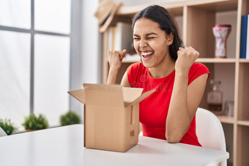 Young brazilian woman looking inside cardboard box celebrating surprised and amazed for success with arms raised and eyes closed. Young brazilian woman looking inside cardboard box celebrating surprised and amazed for success with arms raised and eyes closed