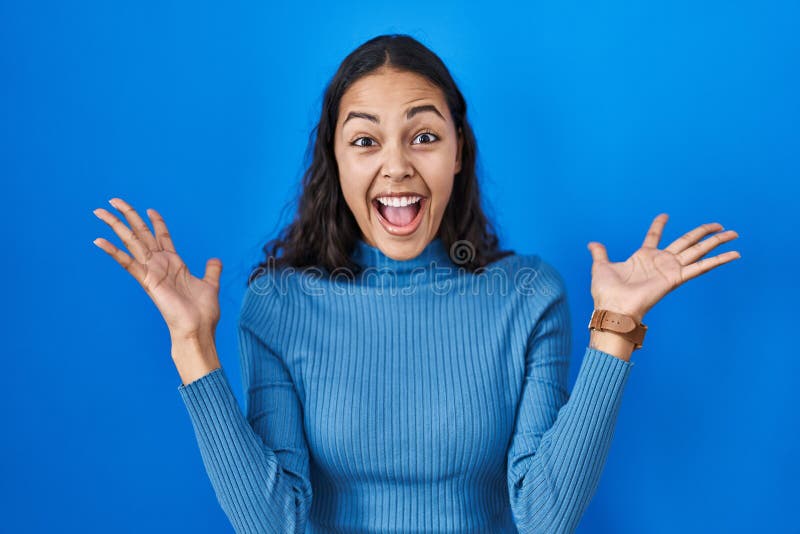 Young brazilian woman standing over blue isolated background celebrating crazy and amazed for success with arms raised and open eyes screaming excited. winner concept. Young brazilian woman standing over blue isolated background celebrating crazy and amazed for success with arms raised and open eyes screaming excited. winner concept