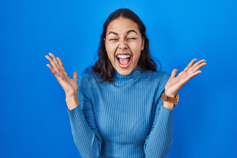 Young brazilian woman standing over blue isolated background celebrating mad and crazy for success with arms raised and closed eyes screaming excited. winner concept. Young brazilian woman standing over blue isolated background celebrating mad and crazy for success with arms raised and closed eyes screaming excited. winner concept