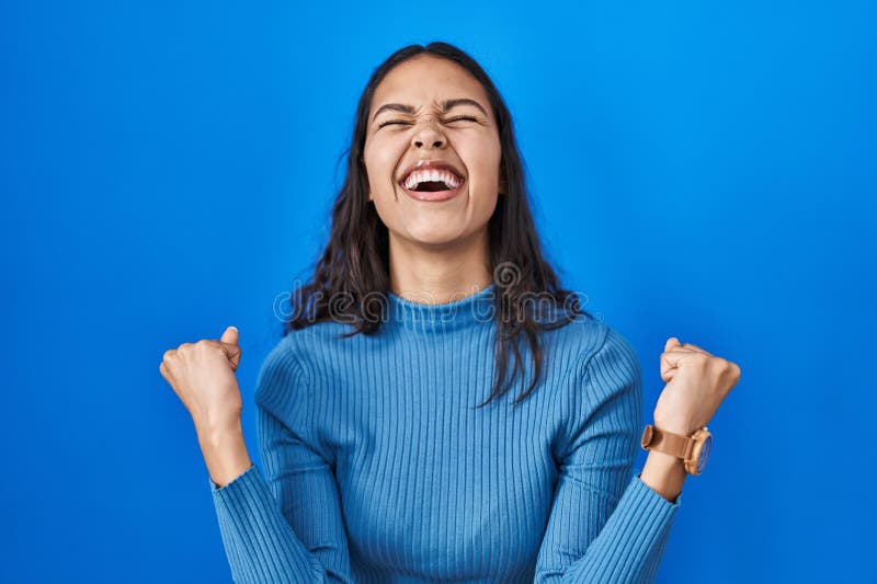 Young brazilian woman standing over blue isolated background celebrating surprised and amazed for success with arms raised and eyes closed. winner concept. Young brazilian woman standing over blue isolated background celebrating surprised and amazed for success with arms raised and eyes closed. winner concept