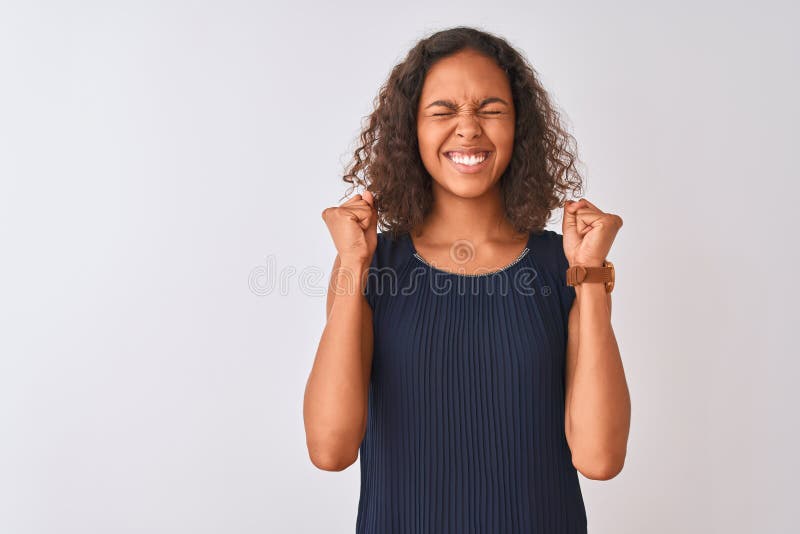 Young brazilian woman wearing blue dress standing over isolated white background excited for success with arms raised and eyes closed celebrating victory smiling. Winner concept. Young brazilian woman wearing blue dress standing over isolated white background excited for success with arms raised and eyes closed celebrating victory smiling. Winner concept