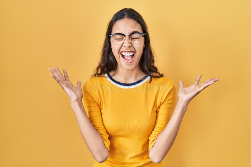 Young brazilian woman wearing glasses over yellow background celebrating mad and crazy for success with arms raised and closed eyes screaming excited. winner concept. Young brazilian woman wearing glasses over yellow background celebrating mad and crazy for success with arms raised and closed eyes screaming excited. winner concept