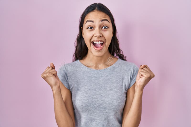 Young brazilian woman wearing casual t shirt over pink background celebrating surprised and amazed for success with arms raised and open eyes. winner concept. Young brazilian woman wearing casual t shirt over pink background celebrating surprised and amazed for success with arms raised and open eyes. winner concept