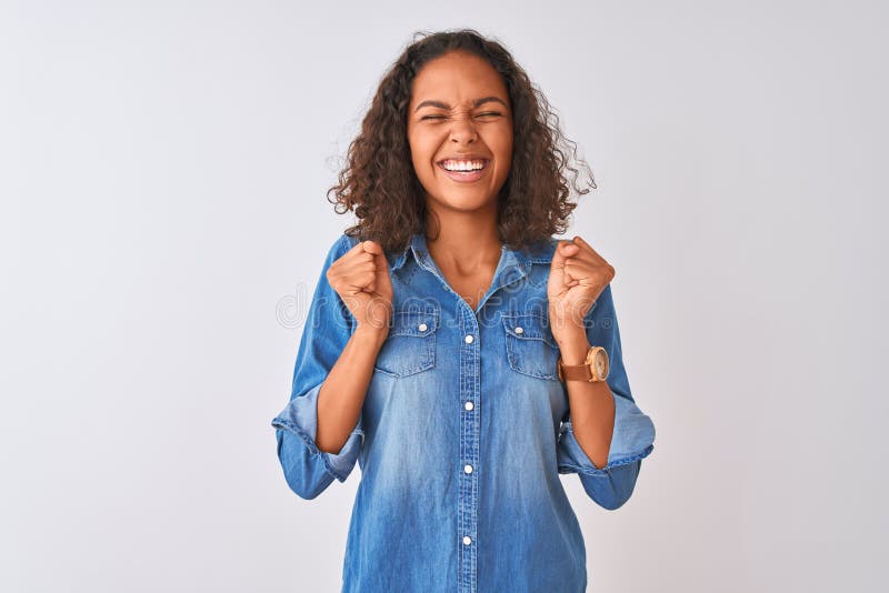 Young brazilian woman wearing denim shirt standing over isolated white background excited for success with arms raised and eyes closed celebrating victory smiling. Winner concept. Young brazilian woman wearing denim shirt standing over isolated white background excited for success with arms raised and eyes closed celebrating victory smiling. Winner concept