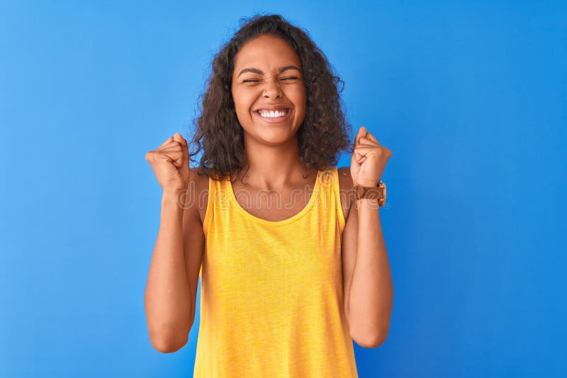 Young brazilian woman wearing yellow t-shirt standing over isolated blue background excited for success with arms raised and eyes closed celebrating victory smiling. Winner concept. Young brazilian woman wearing yellow t-shirt standing over isolated blue background excited for success with arms raised and eyes closed celebrating victory smiling. Winner concept