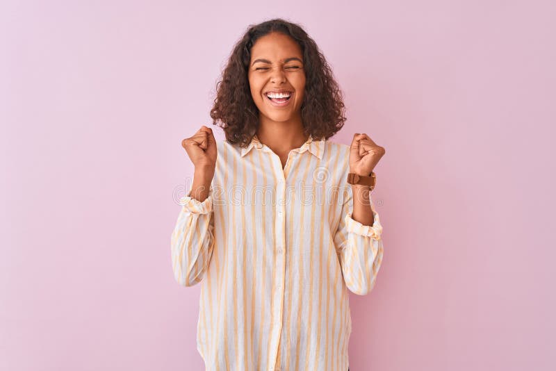 Young brazilian woman wearing striped shirt standing over isolated pink background excited for success with arms raised and eyes closed celebrating victory smiling. Winner concept. Young brazilian woman wearing striped shirt standing over isolated pink background excited for success with arms raised and eyes closed celebrating victory smiling. Winner concept