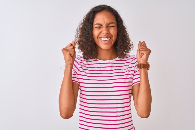 Young brazilian woman wearing pink striped t-shirt standing over isolated white background excited for success with arms raised and eyes closed celebrating victory smiling. Winner concept. Young brazilian woman wearing pink striped t-shirt standing over isolated white background excited for success with arms raised and eyes closed celebrating victory smiling. Winner concept