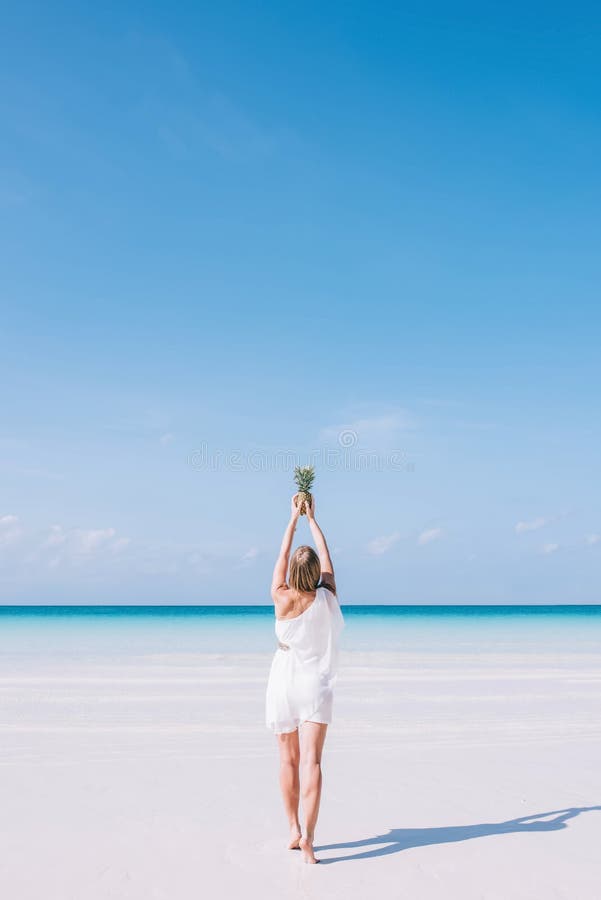 A young European long hair woman stands on a white sandy beach by the ocean. A girl in a white dress is holding a pineapple in her hands. Women is looking at the tropical turquois sea. Summer vacation concept. A young European long hair woman stands on a white sandy beach by the ocean. A girl in a white dress is holding a pineapple in her hands. Women is looking at the tropical turquois sea. Summer vacation concept.