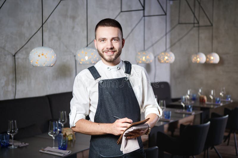 Een jonge, glimlachende kelner in een restaurant, die zich naast de lijsten met een glas wijn bevinden Gekleed in een schort, een stock afbeeldingen Een jonge, glimlachende kelner in een restaurant, die zich naast de lijsten met een glas wijn bevinden Gekleed in een schort, een stock afbeeldingen
