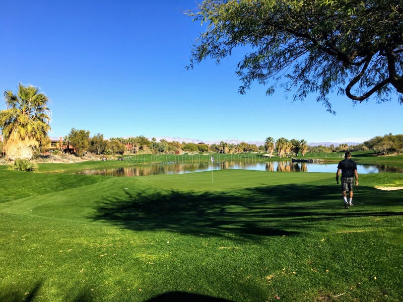 Een jonge mannelijke golfspeler die naar green op een pari 4 lopen die door water op een golfcursus wordt omringd in de woestijno stock afbeelding Een jonge mannelijke golfspeler die naar green op een pari 4 lopen die door water op een golfcursus wordt omringd in de woestijno stock afbeelding