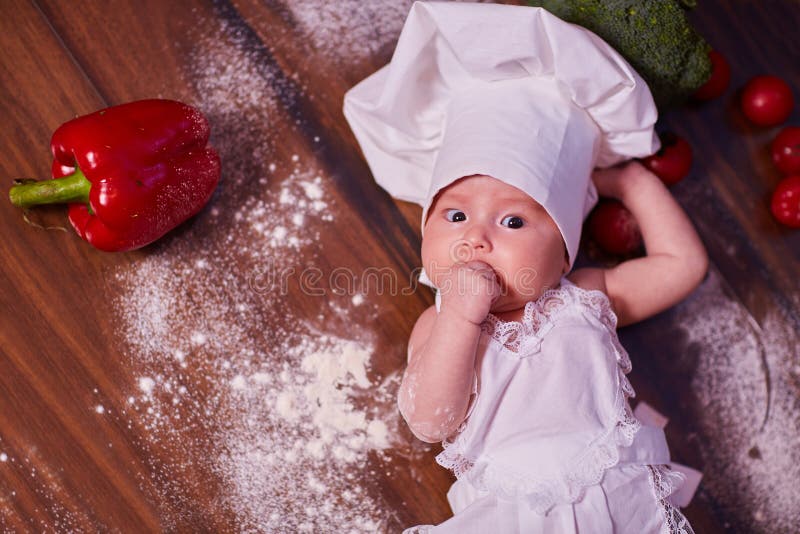 Een kind, baby, meisje, ligt op de keukenlijst, in GLB van een kok en in een schort, in witte sokken, naast bloem, groenten, pepp stock foto's Een kind, baby, meisje, ligt op de keukenlijst, in GLB van een kok en in een schort, in witte sokken, naast bloem, groenten, pepp stock foto's