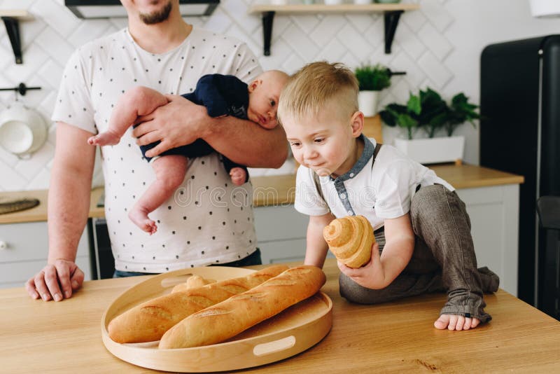 Een kleine jongenszitting op de keukenlijst en holding een croissant met zijn vader die een baby achter hem houden stock fotografie Een kleine jongenszitting op de keukenlijst en holding een croissant met zijn vader die een baby achter hem houden stock fotografie