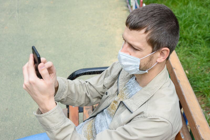 A man in a protective medical mask sitting on a bench in the park communicates by cell  phone. A man in a protective medical mask sitting on a bench in the park communicates by cell  phone