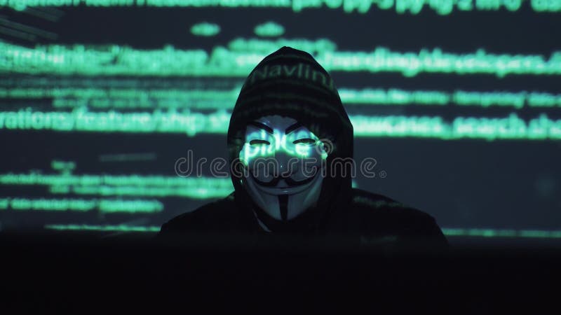 Een mannelijke rover in het maskerwerk aangaande een computer in een donkere ruimte de computercode wordt overdacht zijn gezicht